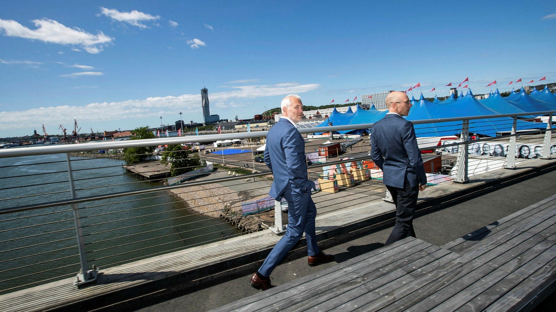 Gående på Hisingsbron över Göta älv: Till vänster Jonas Attenius (S), ordförande i kommunstyrelsen, till höger Henrik Einarsson, etableringsansvarig vid Business Region Göteborg. Det blå tältet i Frihamnen framför dem är platsen där Polestar ska bygga sitt nya huvudkontor. Karlatornet syns i bakgrunden.